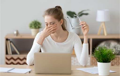 A young woman sitting at a table with her laptop taking a break and rubbing her eyes while she holds her glasses in her other hand.