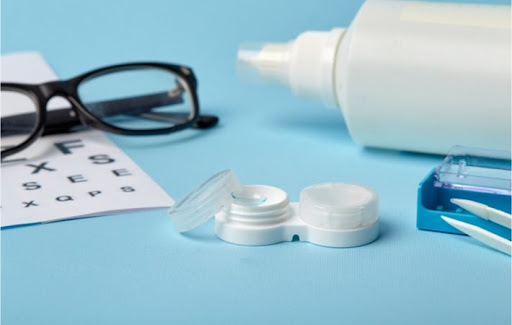 Blue surface with several optical items on it including a contact lens case with one part open and a lens visible, pair of glasses on top of an eye chart, bottle of contact lens solution laying sideways, and forceps laying next to their open case.
