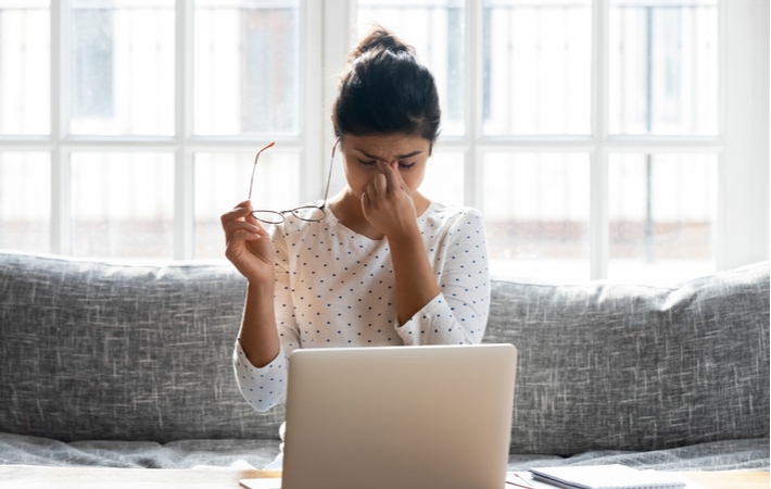 A woman sitting on her couch using her laptop, holding her glasses in one hand and her rubbing her eyes with the other as she suffers from discomfort due to digital eye strain