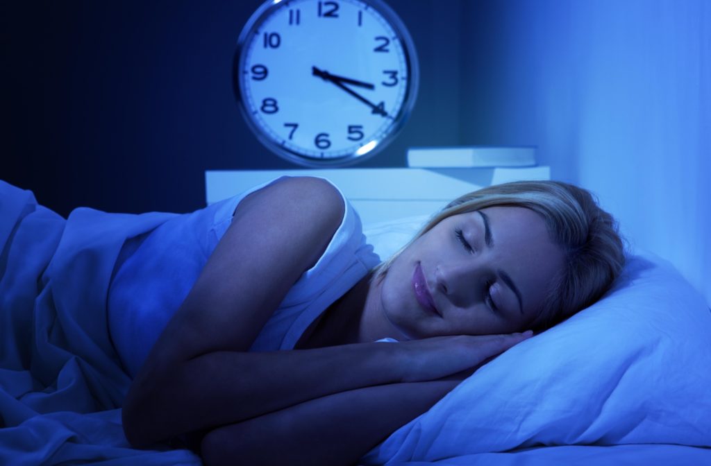 A woman sleeping on her side at night with a large clock behind her reading 3:20am