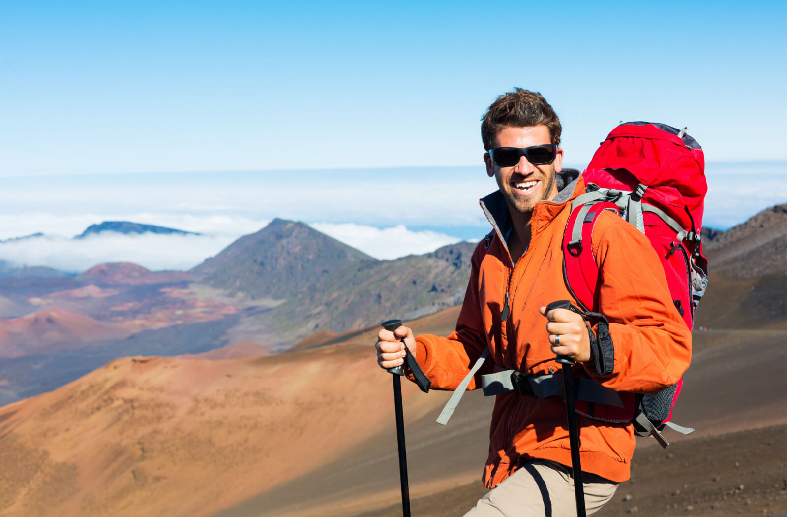 A man on top of a mountain wearing sporty sunglasses and a backpack while hiking with poles