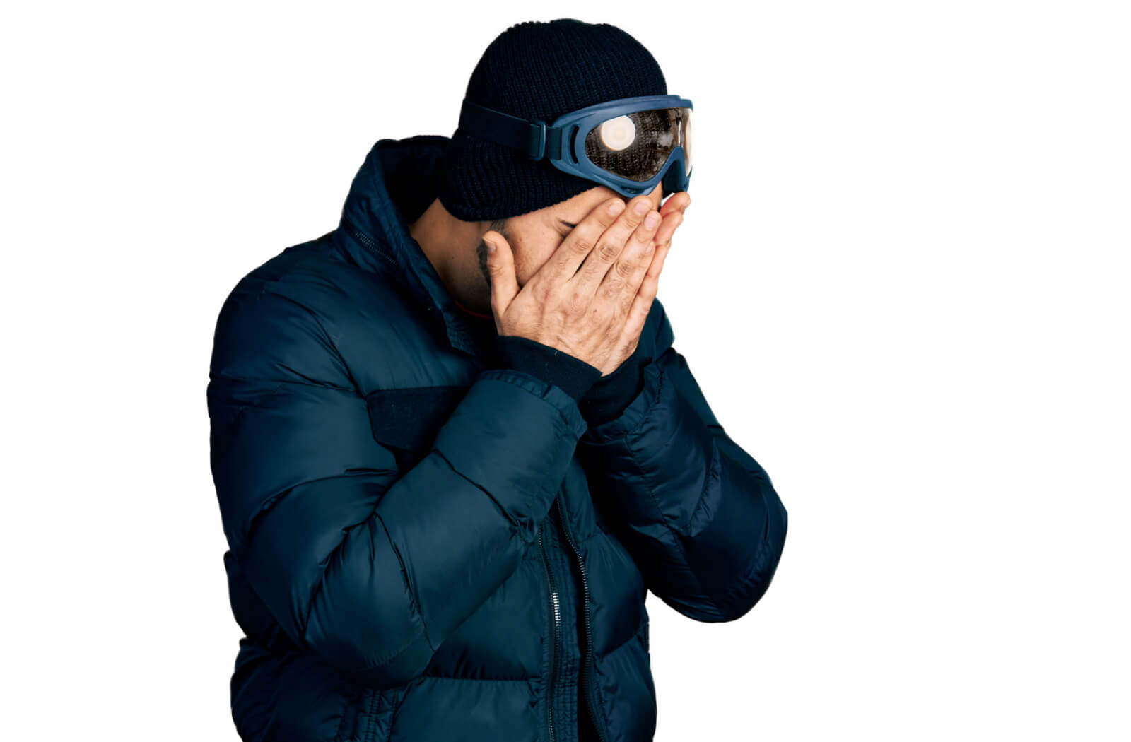 A man in a winter coat and snow goggles against a white background covering his eyes with both hands