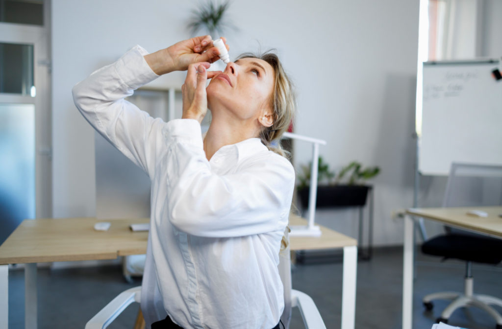 A professional-looking young woman in an office space applying eye drops on her right eye.