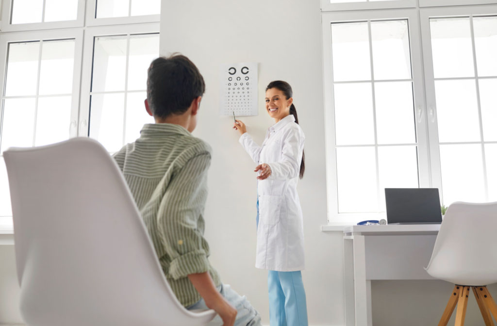 View from behind a child's head as he sits in a chair and looks ahead. His eye doctor is smiling and pointing to a Landolt C eye chart on the wall.