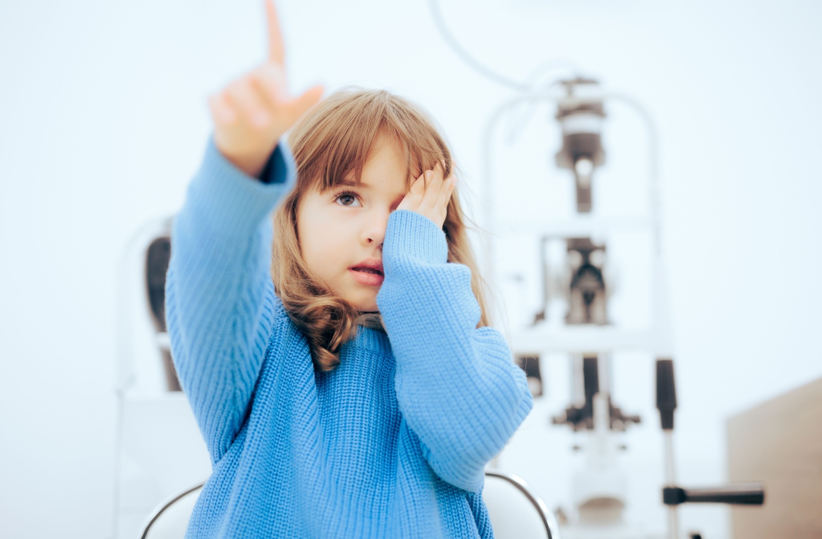 A little girl in a blue sweater covering one eye and pointing during an eye exam to diagnose her myopia.