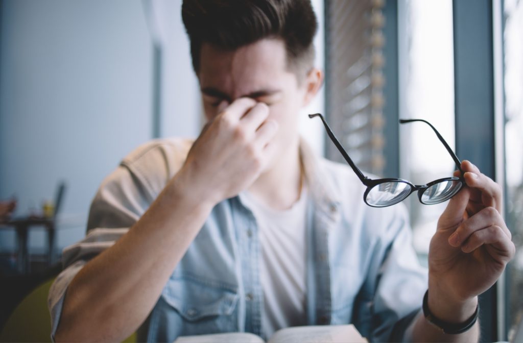 A slightly out-of-focus image of a young adult taking off their glasses to rub their eyes in frustration from blurry vision caused by dry eyes.