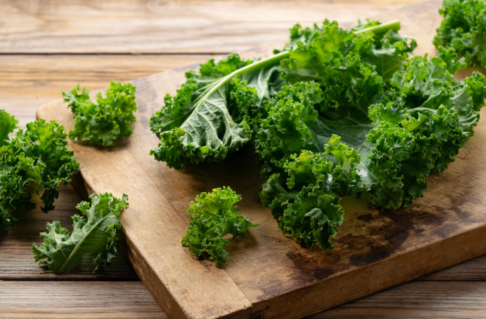 Pieces of kale lay on top of a cutting board sitting on a wooden surface.