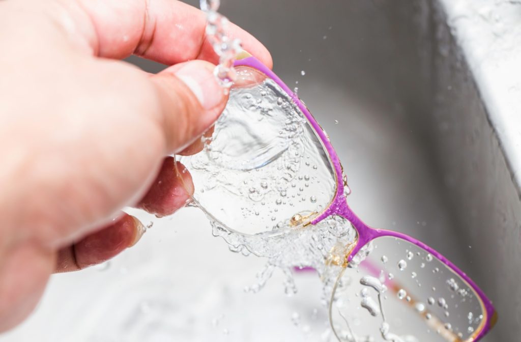 Someone’s hand holds a pair of pink framed eyeglasses under running tap water as an initial step to cleaning them