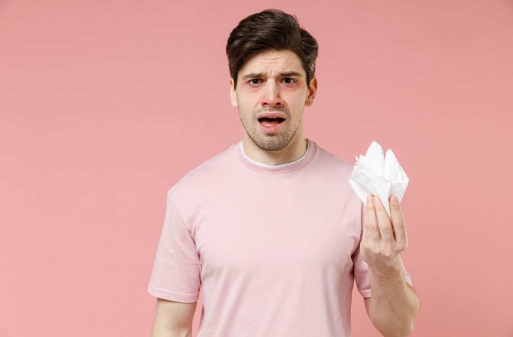 Against a pink background, an adult in a pink shirt stands with red, swollen eyes and a tissue in hand