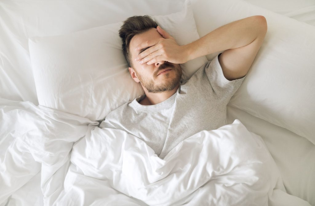 A person waking up in bed covers their eyes due to pain.