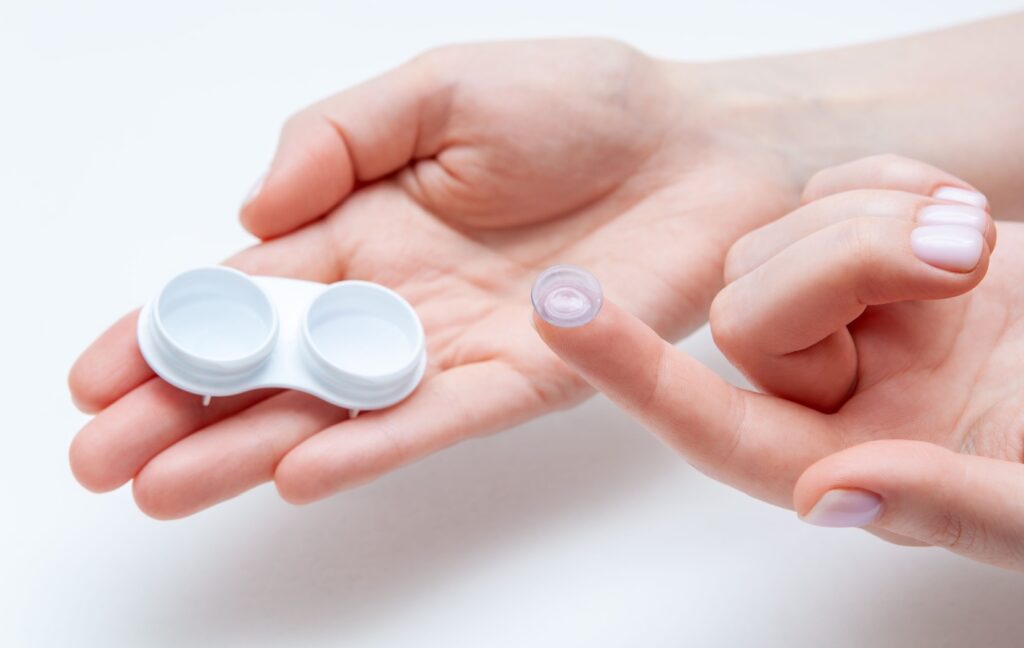 A person holds an open, white contact lens case in one hand while they hold up a contact lens with their other hand.