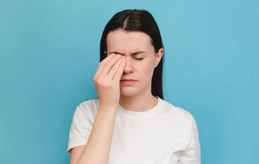 A person wearing a white shirt has sore, burning eyes. They squint as they rub their left eye with their hand for relief.