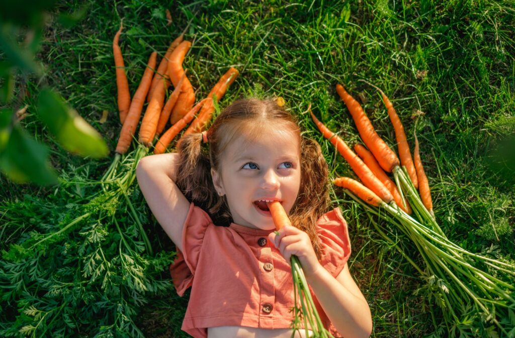 A child snacking on a carrot while lying on grass, surrounded by a handful of carrots.