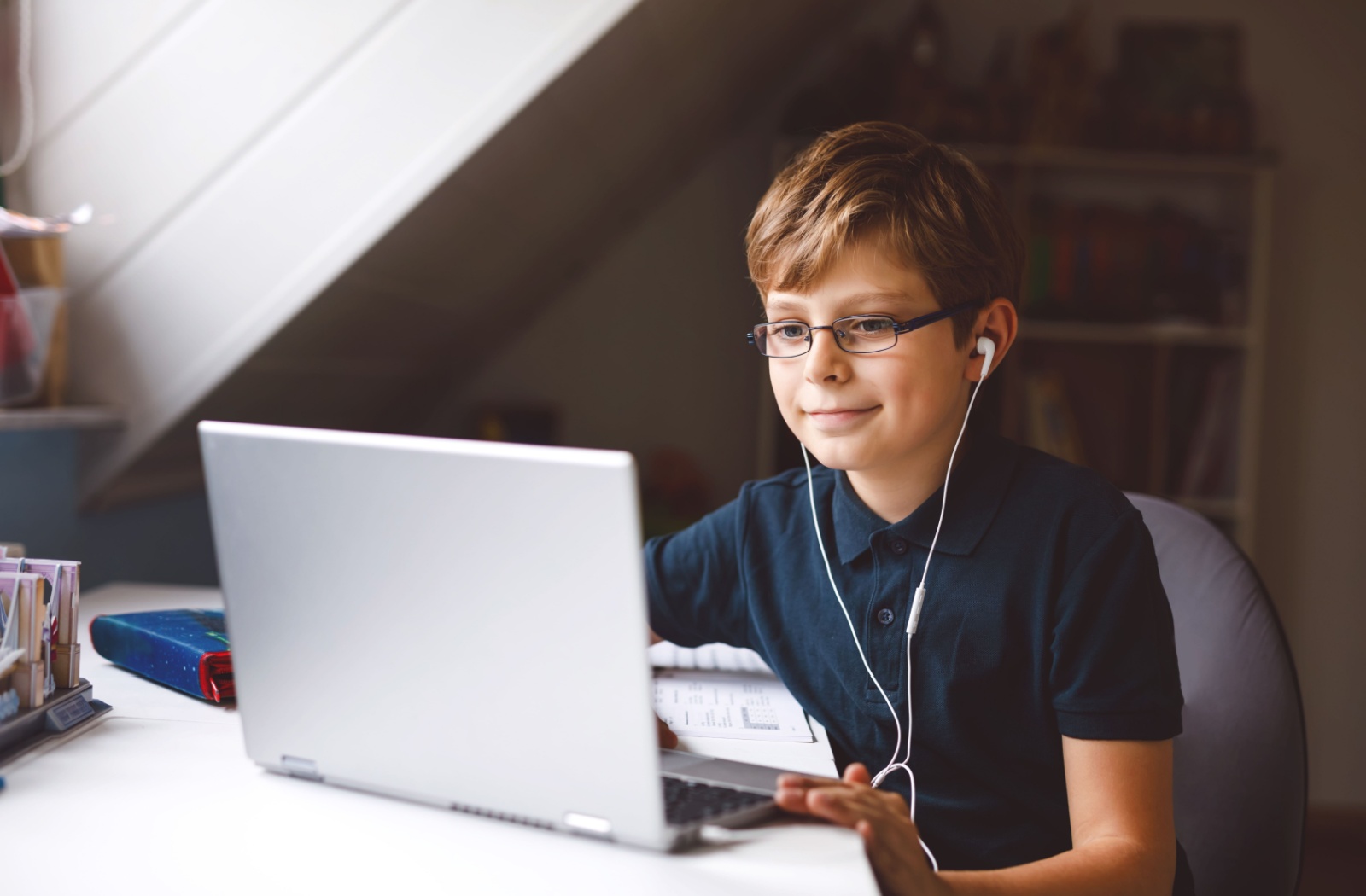 Young smiling boy with glasses wearing earphones and doing homework on a laptop.